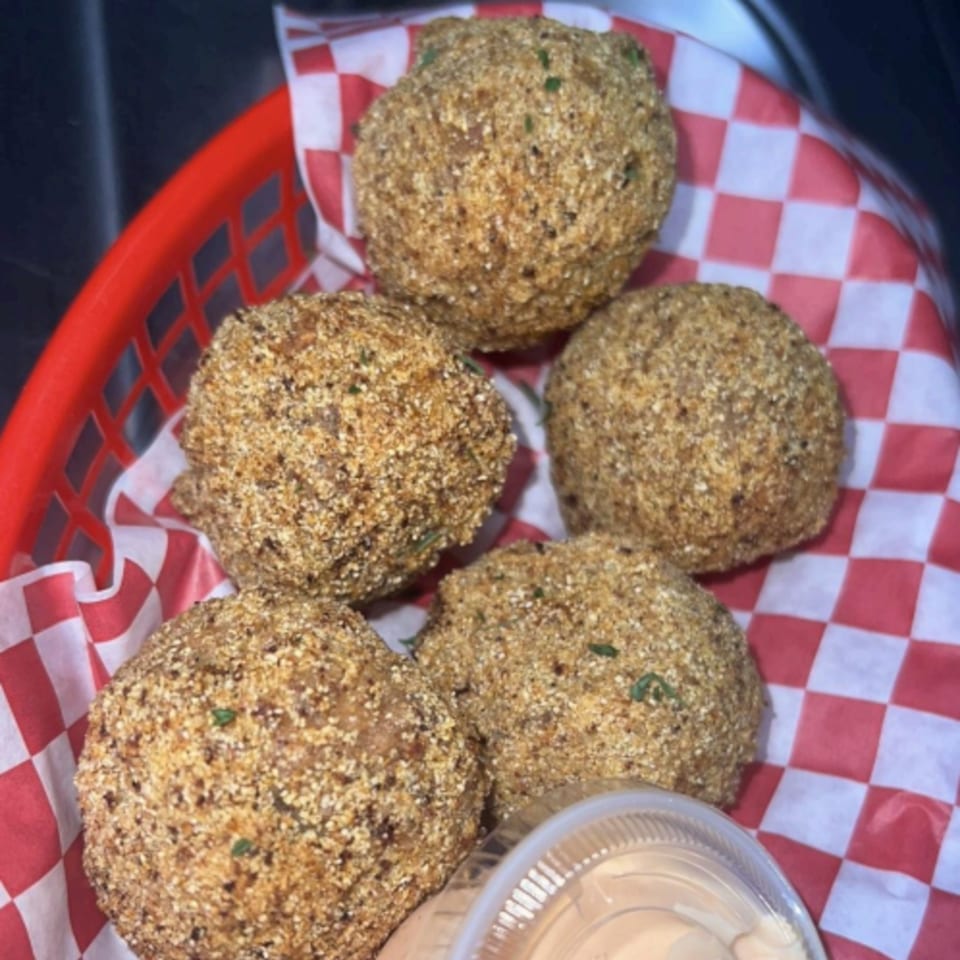 Five crispy, breaded seafood balls in a red basket with checkered paper, alongside a dipping sauce.